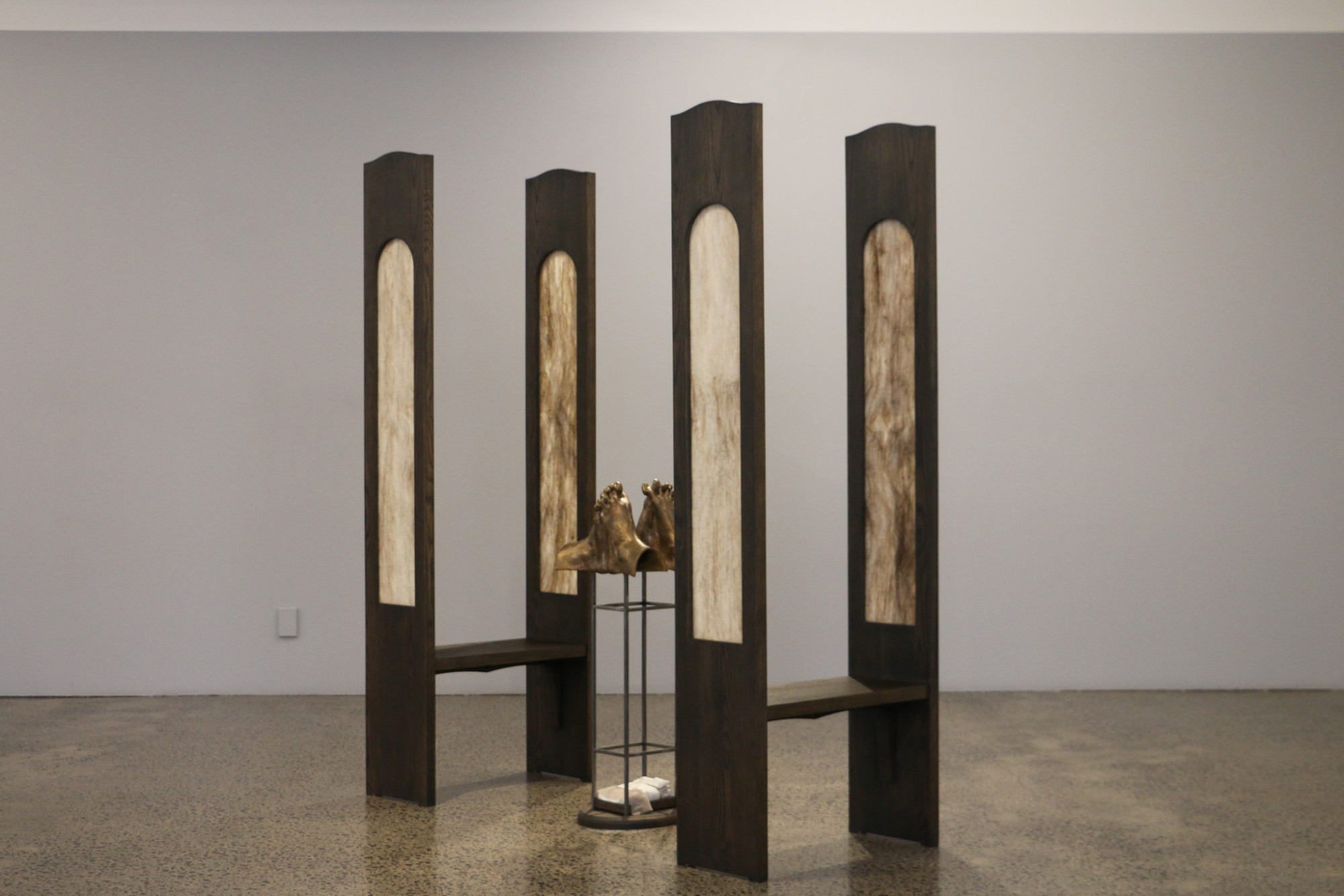 Two pew like seats face each other with a metal structure in between. The structure holds up two sets of feet with intertwined toes cast in bronze.