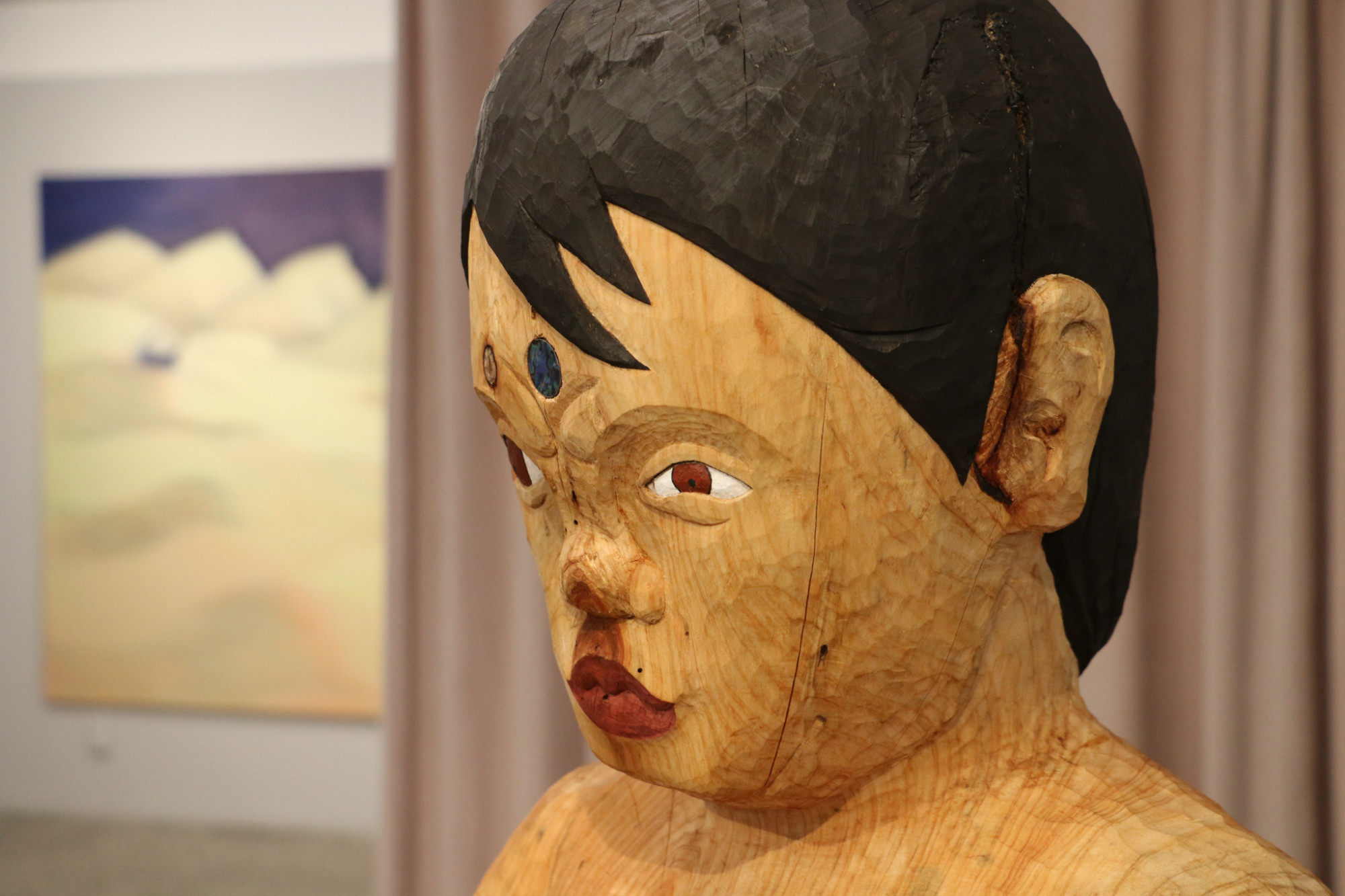 A close up of a carved tekoteko or figure's face with painted features and paua sphere on her forehead. The figure has one eye open and one is partially closed, forming a focused expression.