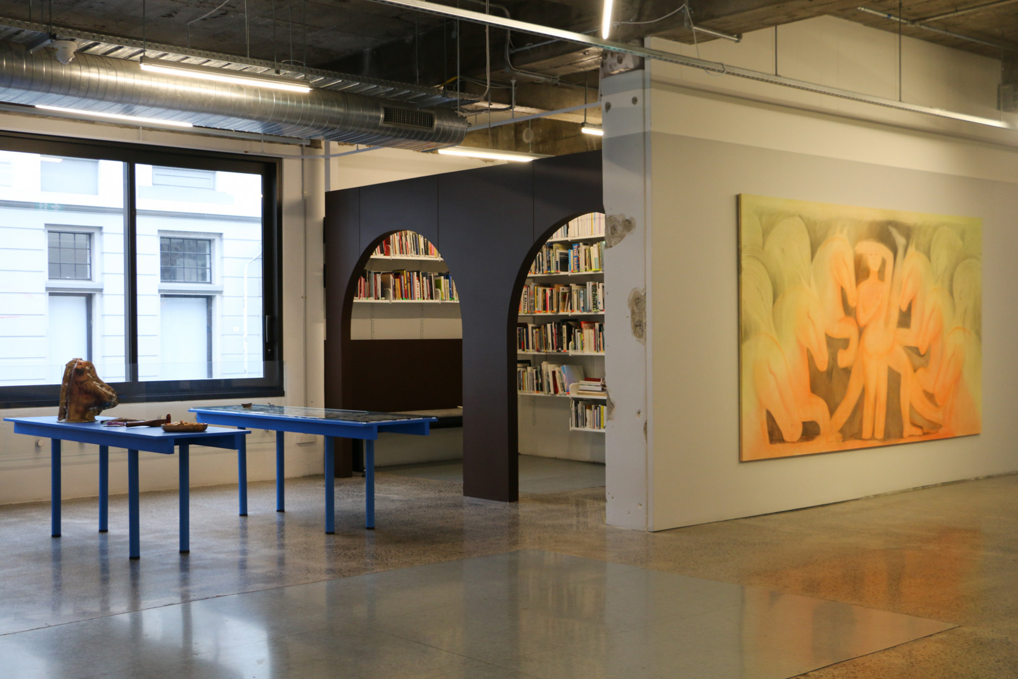 A gallery space with a large painting on one wall, blue long tables with objects on them and a library space in the background.