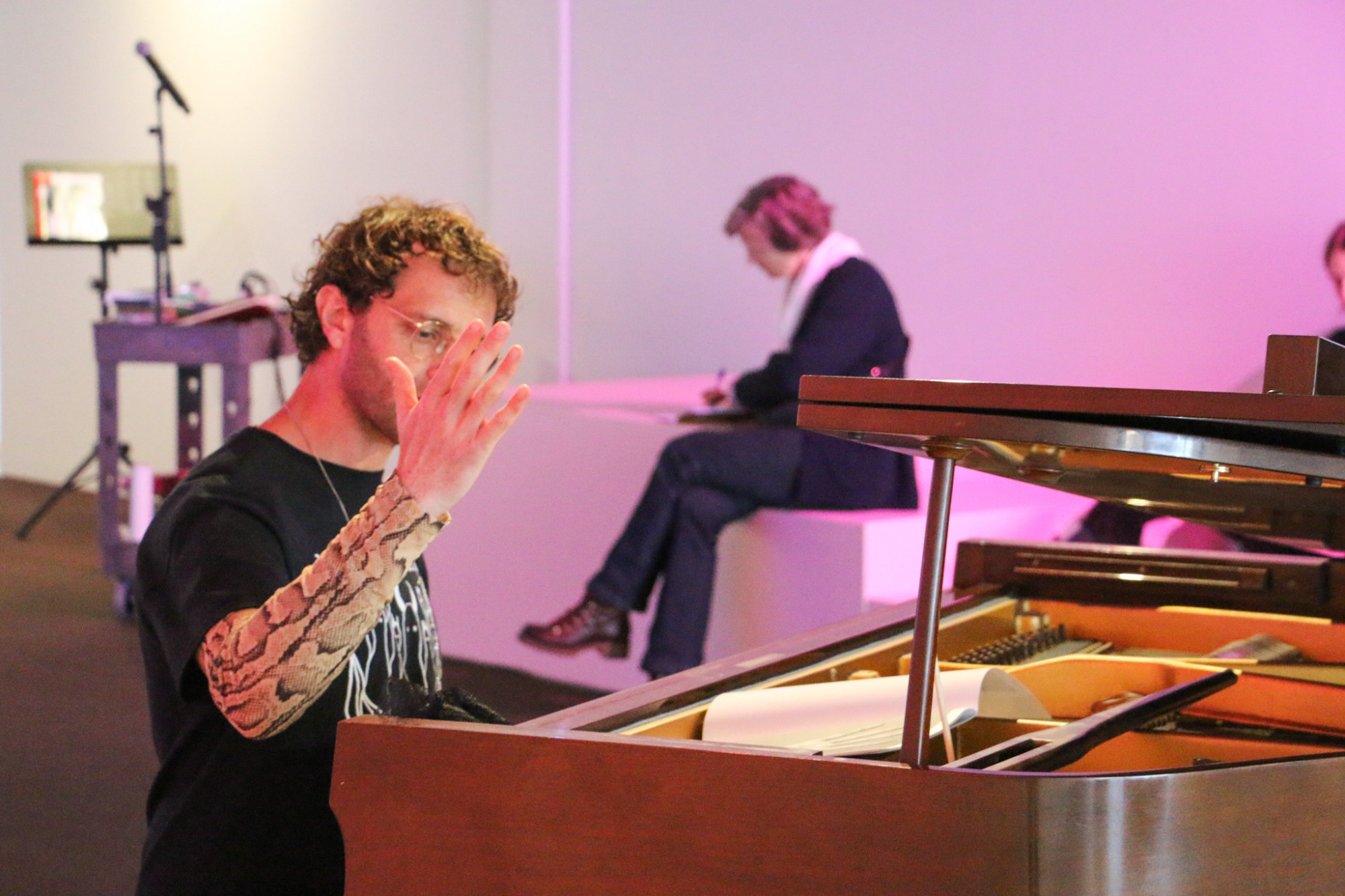 A person is sat at a piano, they are holding their arm upright at a slight angle, a person is seated on the flat staircase behind them.