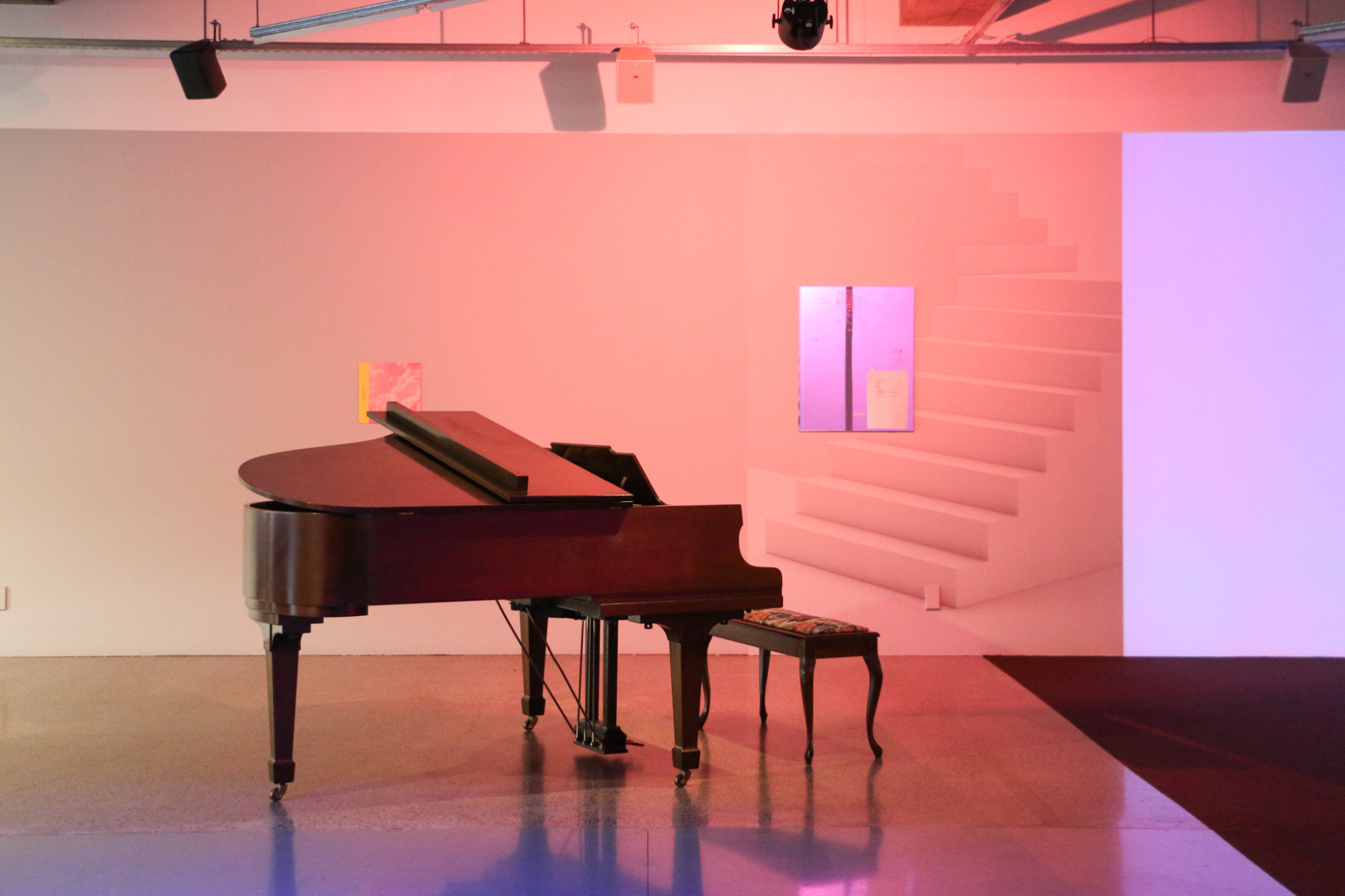 A piano and stool are in a gallery surrounded by artworks and a projection of purple light on the wall.