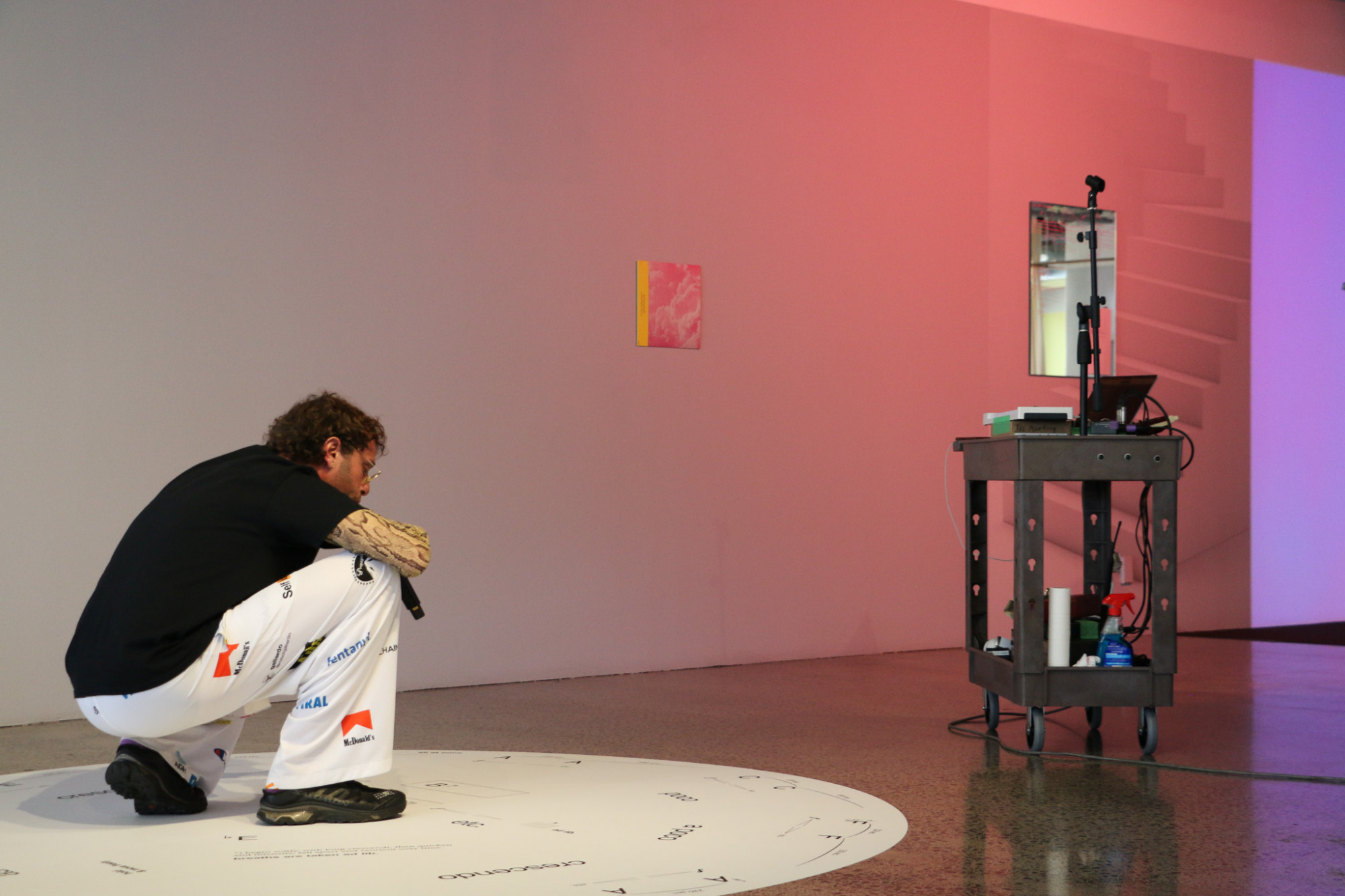 In the gallery a person is crouched down on a circular floor graphic and is holding a microphone  to their mouth, a trolly with technical equipment is near by.