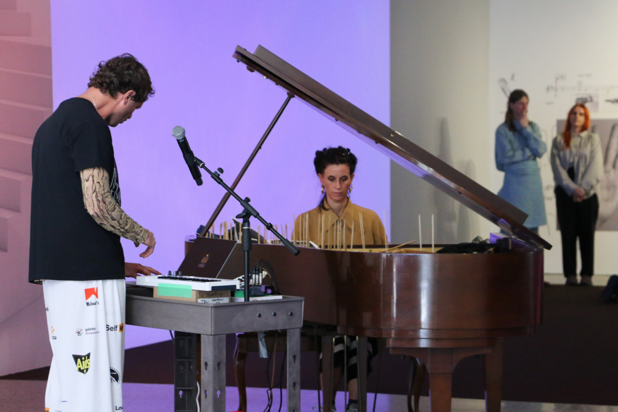 One person is playing a keyboard, a mic stand is in front of them as well as someone seated at a open piano with chopsticks sticking out of it.