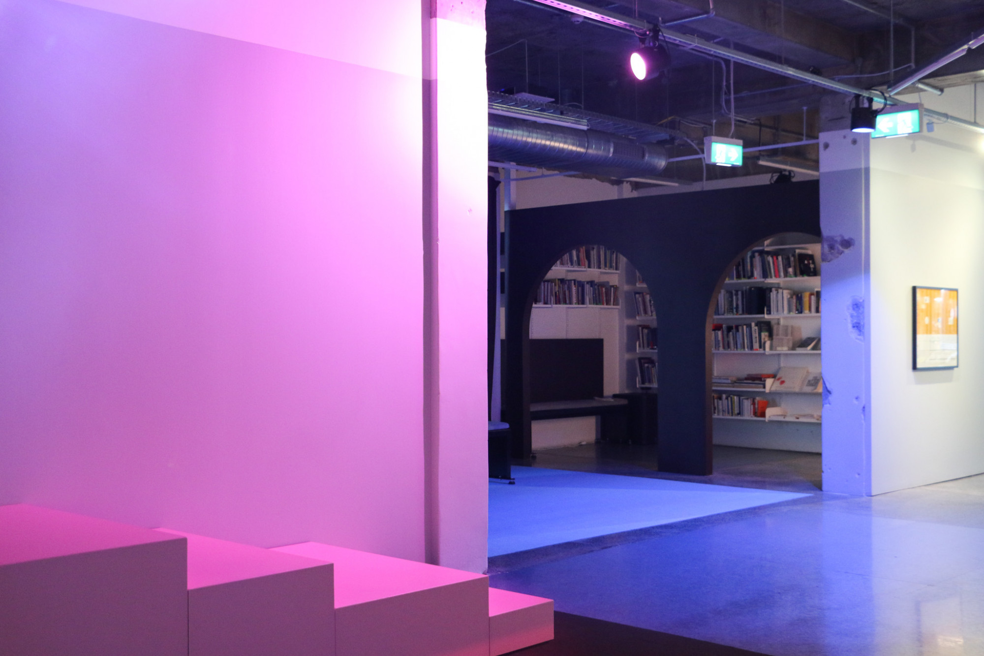 A small flat staircase runs beside a wall with pink light, in the background another room can be seen with  two archways and blue light.