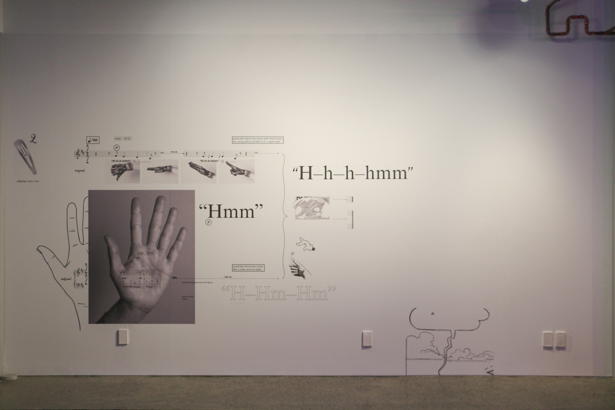 On the gallery wall is large and small text and images and drawings of hands and other symbols and written music.