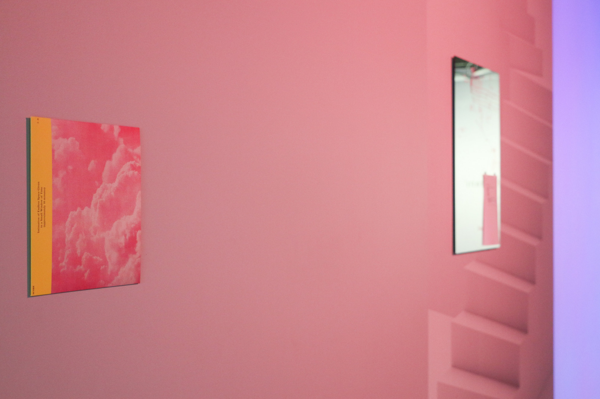 An artwork the size of a record sleeve with text and pink clouds is on the wall, next to a mirror with text and symbols written on it, with a wall decal featuring a staircase behind it all lit by pink light.