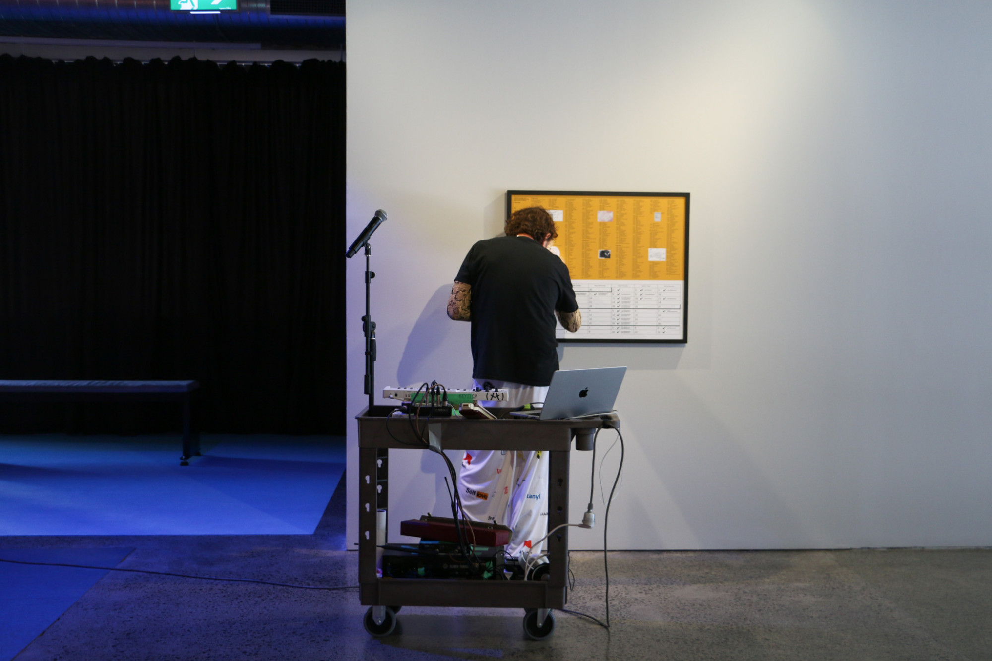 A person stands close to a framed artwork on the wall, next to them is a mic stand and in front of them is a trolley with technical equipment and a laptop.