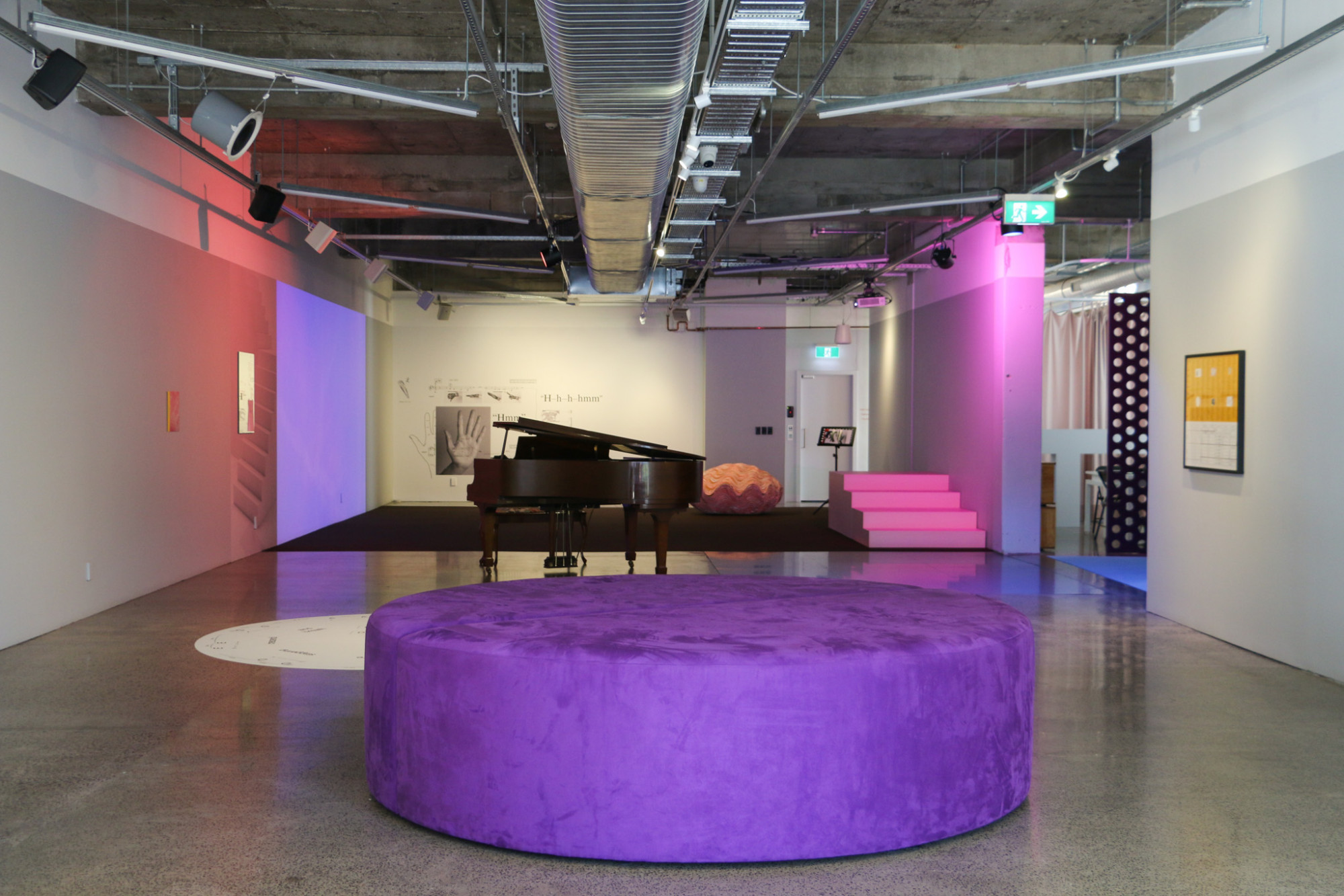 The gallery has a large round purple seat and a piano with artworks on the walls and pink lights.