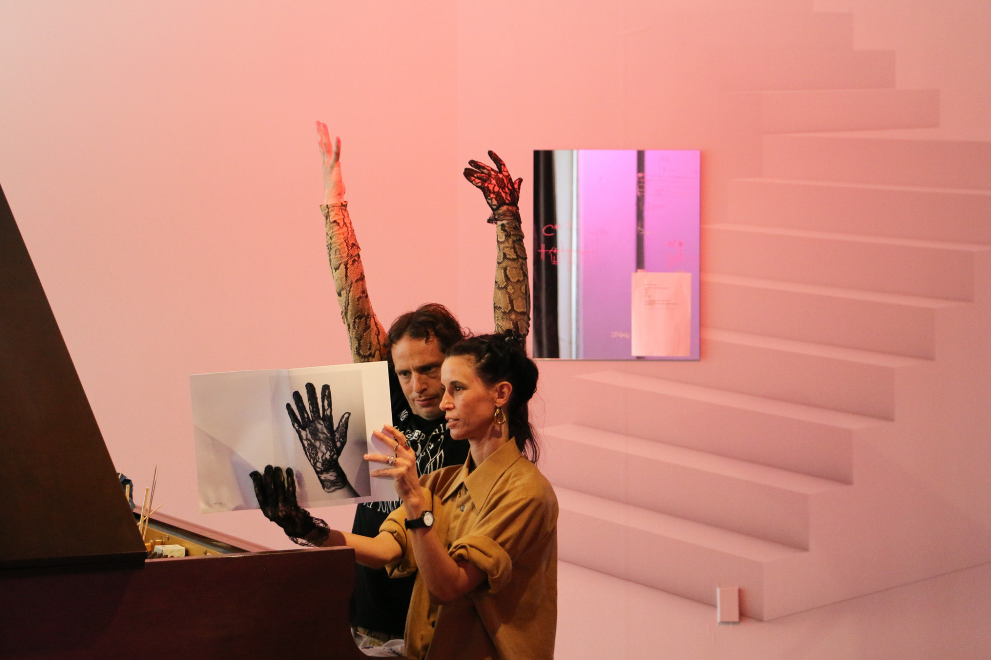 Two people are sat at a piano reading from a booklet with a lace hand on the front which one of them is holding, the other has their hands in the air, both are wearing a lace glove.