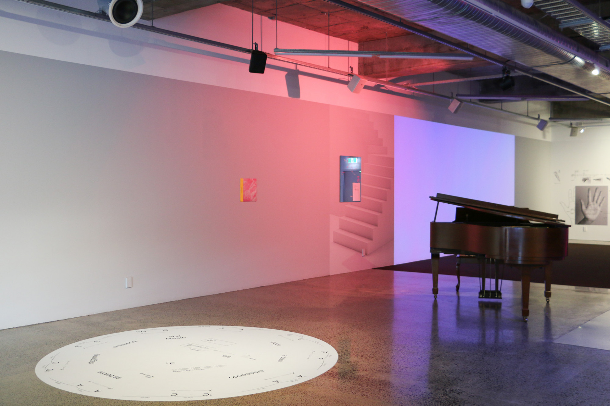 The gallery has a large white circular graphic on the floor which has black text and symbols on it, behind it are a piano and pink light and artworks on the wall.