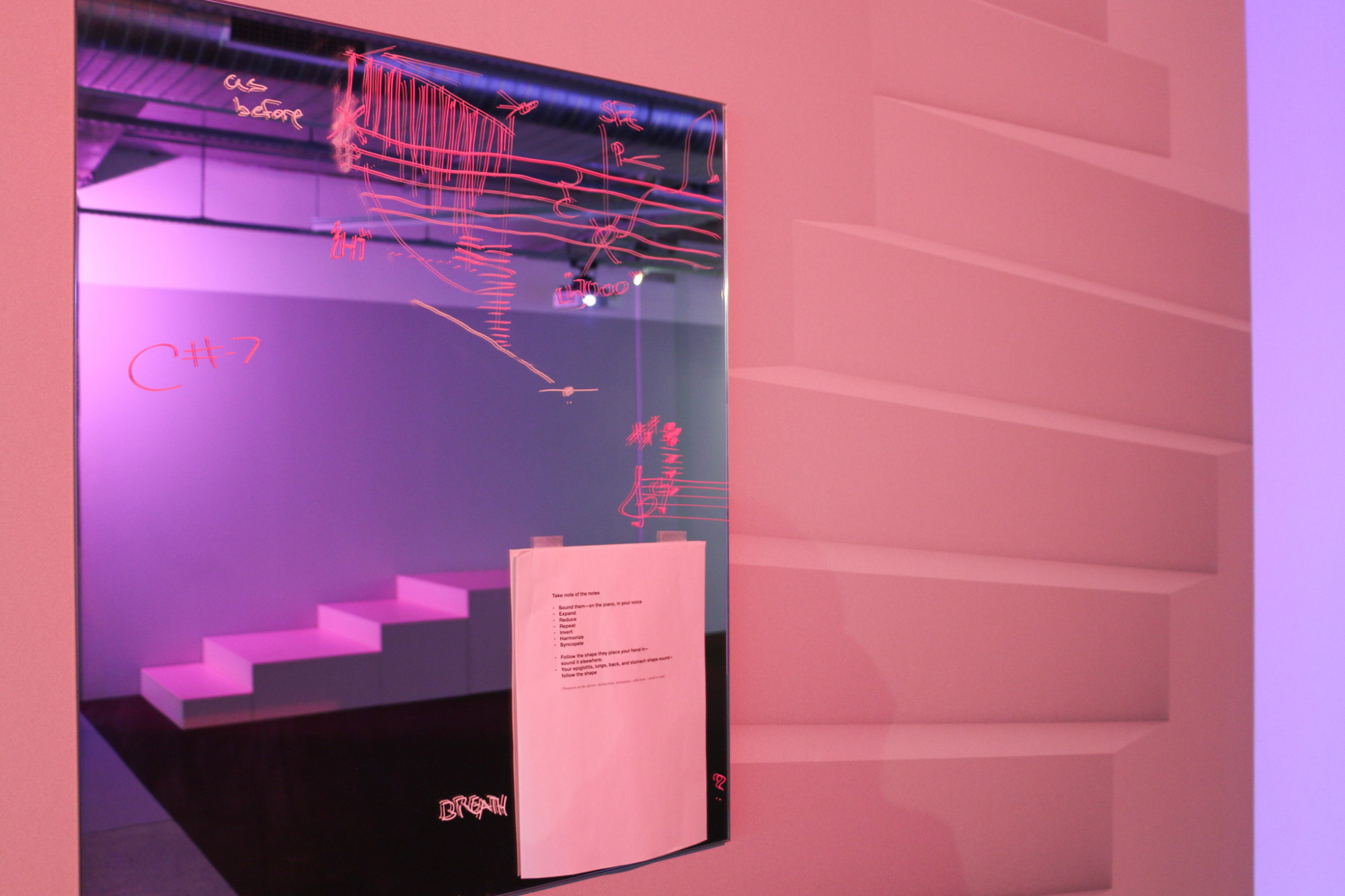 A mirror is hung on the wall, it has pink writing and symbols on it as well as a A4 paper taped with printed instructions, there is a staircase graphic on the wall and stairs are in the reflection of the mirror.