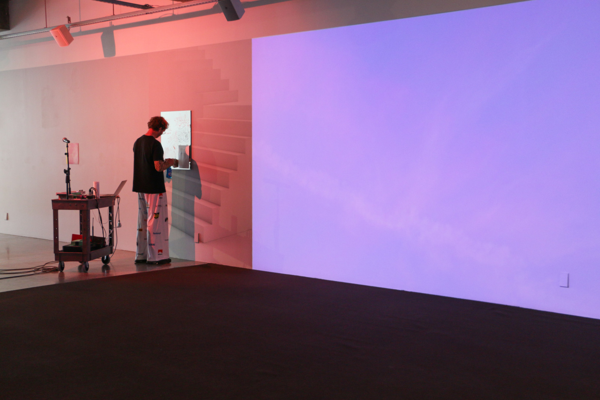 A person is writing on the mirror on the wall, next to them is a large projection of pink and blue clouds and a large dark purple carpet.