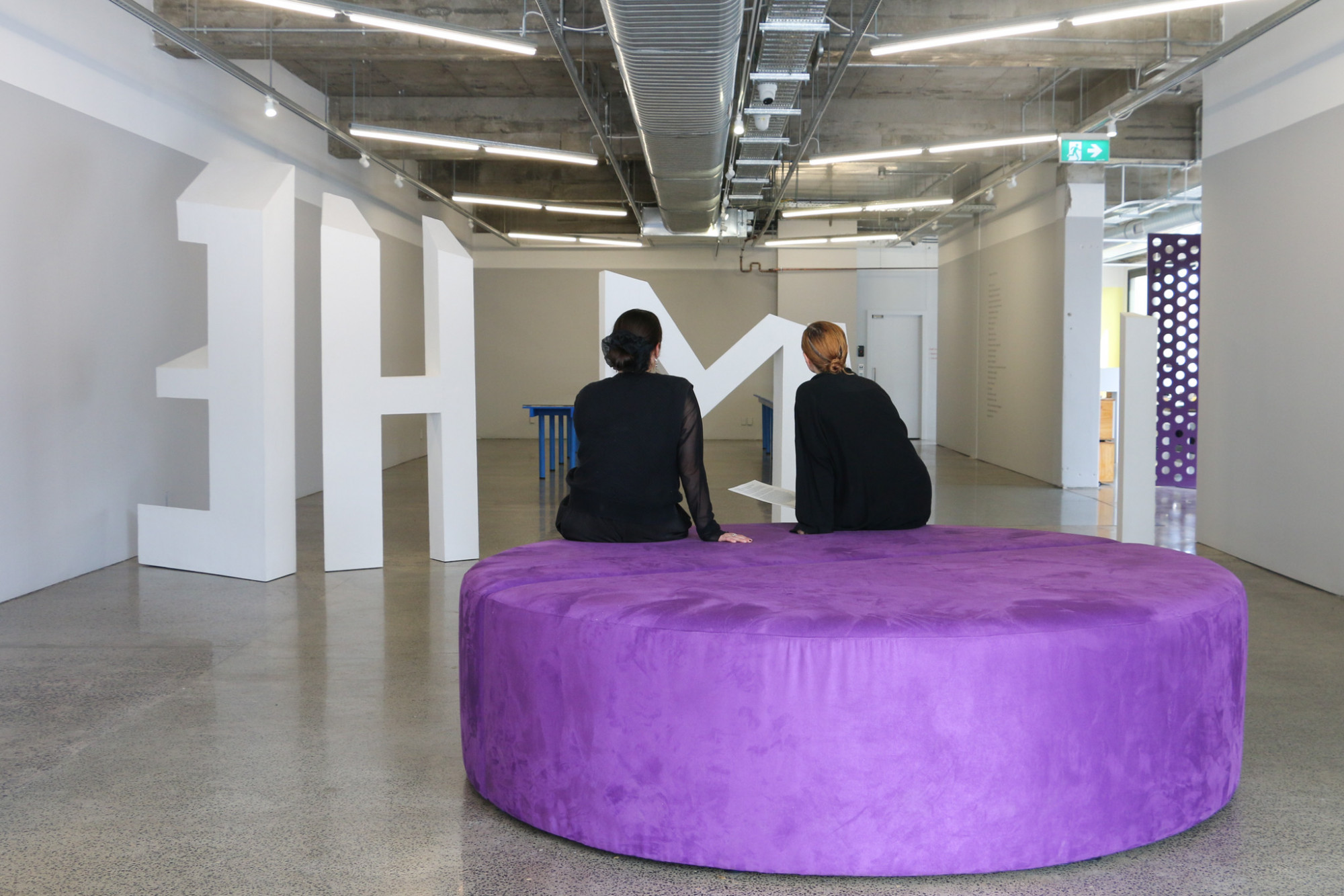 The gallery with a large round purple seat with two people seated on it with their backs to camera. In the background large white sculptural letters spell out, 'I AM HE' they are read backwards from this view point.