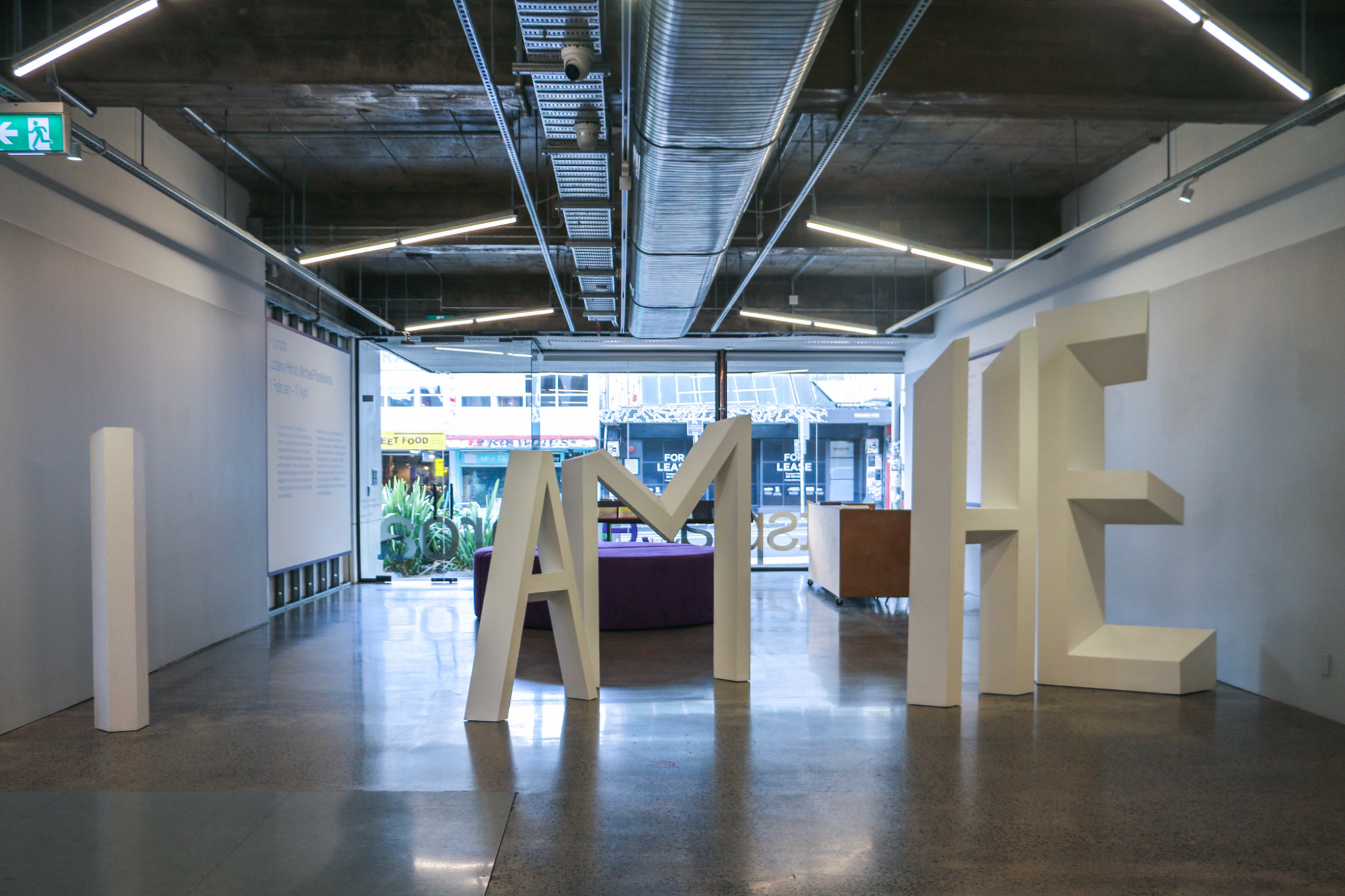 A front view of a larger white sculpture which is positioned across the width of the gallery. It is made up of letters which read 'I AM HE' there is enough space for people to walk between the words. The gallery entrance and foyer is in the background.