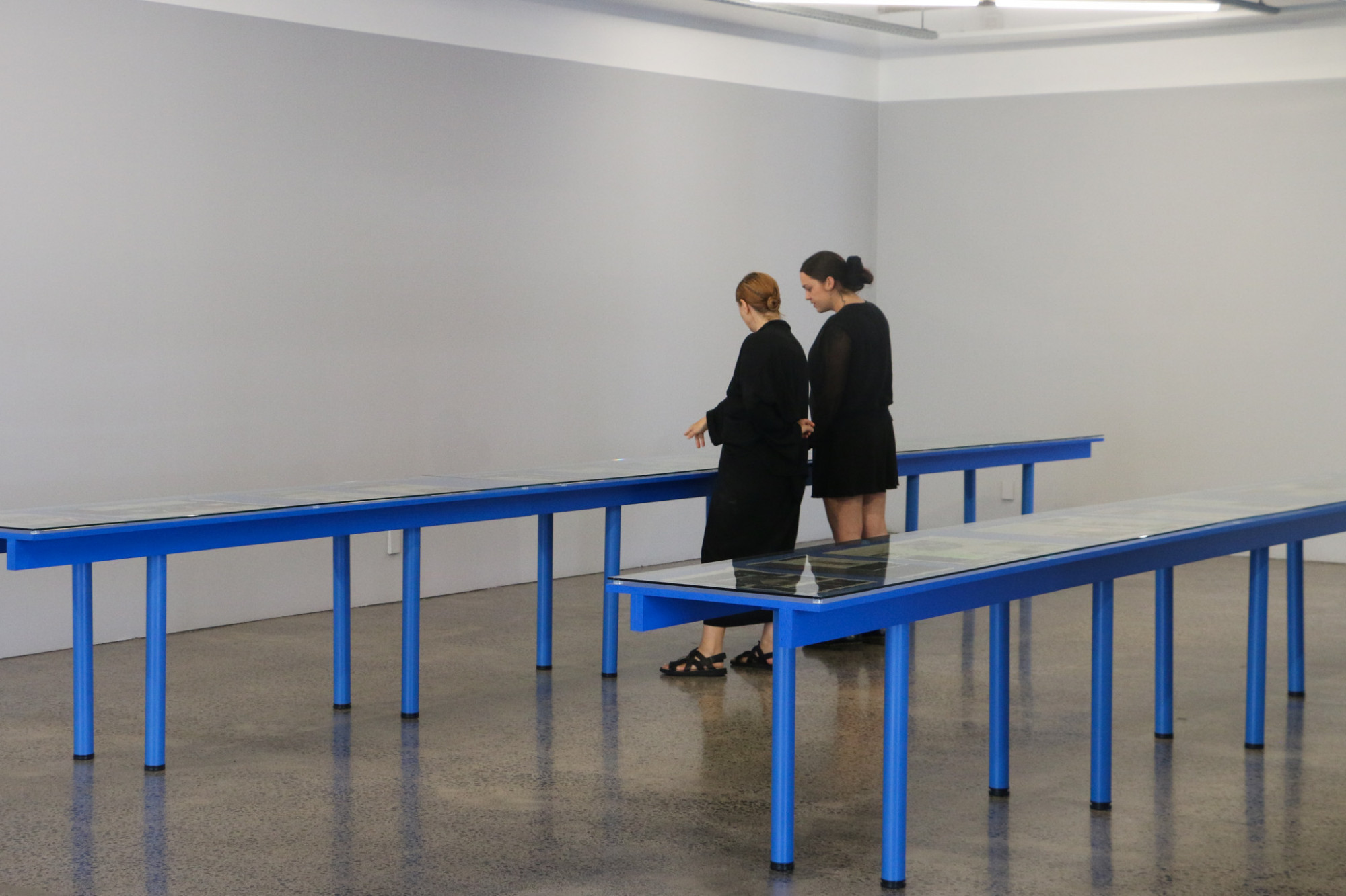 Two long flat vitrines displaying artworks run parallel in the back end of the gallery. They have glass tops and are painted bright blue, two people are looking at the artworks, which are not visible from this distance.