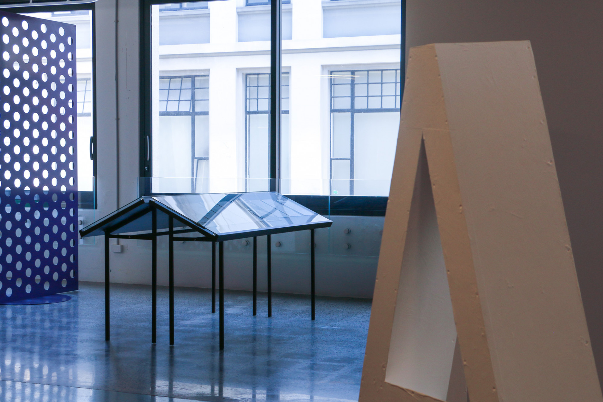 Image shows gallery windows with vtrines painted brown in the background, in the foreground is the top of an 'A' shaped white sculpture.