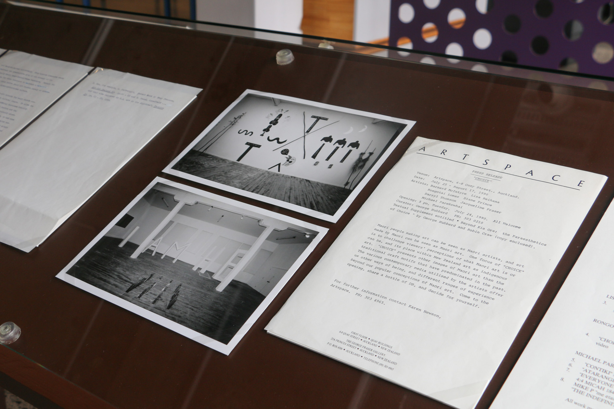 Close up of a vitrine, painted brown with glass on top of some black and white photographs of an exhibition of various sculptures and other documents.