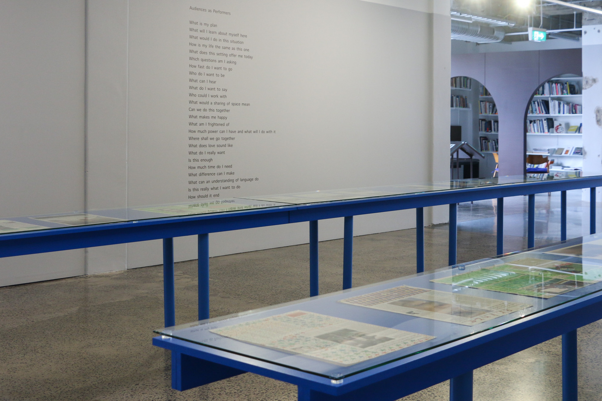 Long blue vitrines displaying newspaper paintings run parallel to each other next to a poem on grey gallery wall, with a room with books and chairs in the distance.