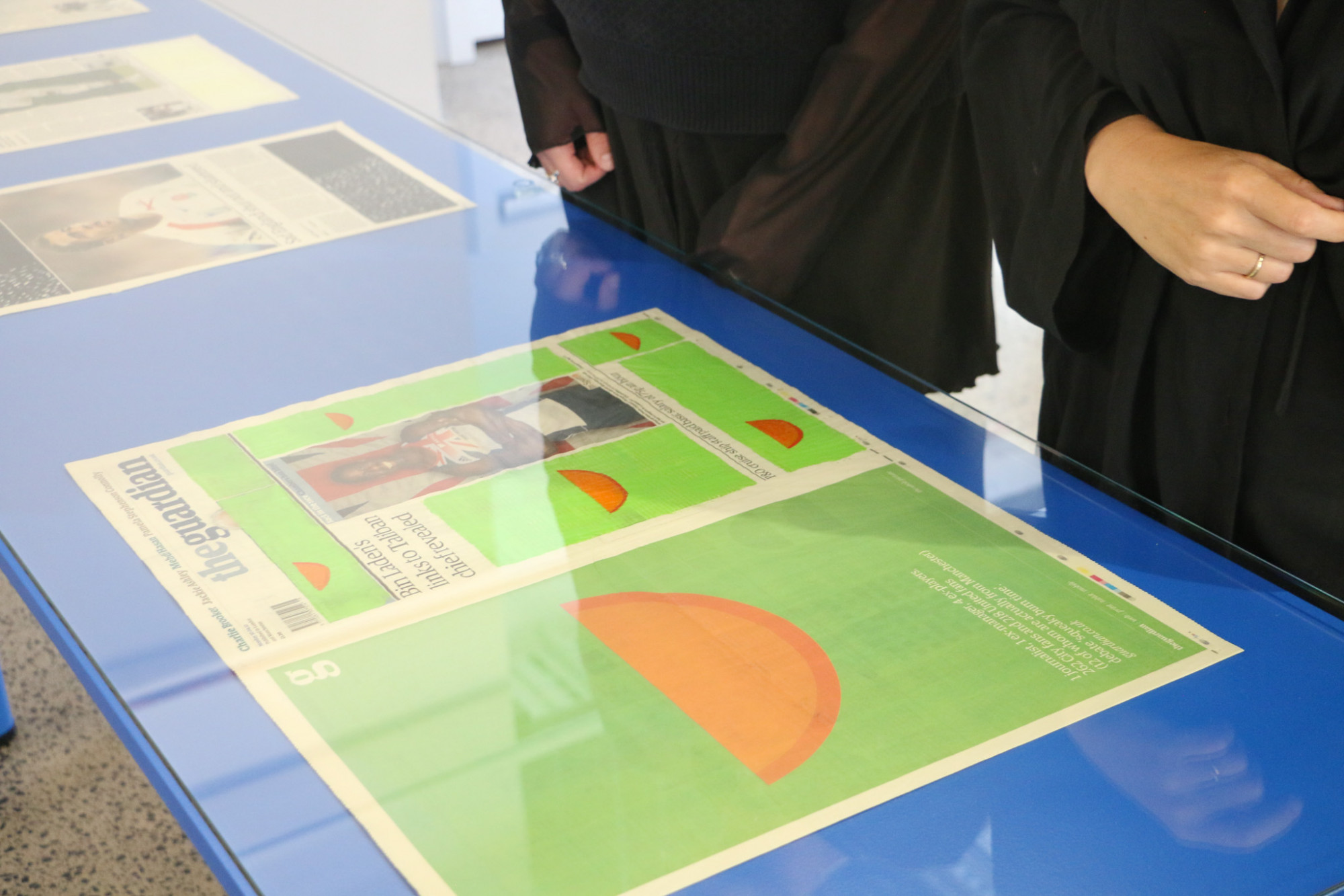 A close up image of the vitrine with newspaper pages with green and orange paintings overlaid, two people's hands and arms are next to the vitrine.