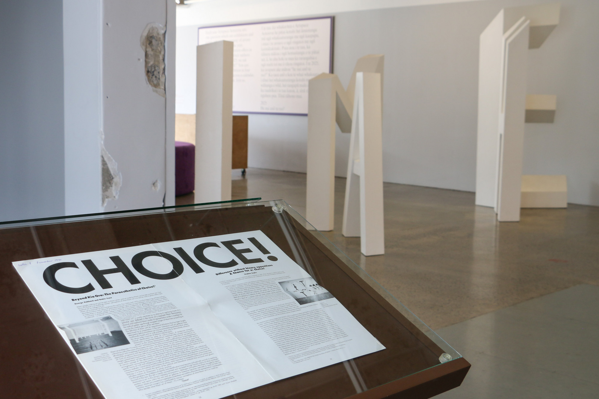 In the foreground  the end of a vitrine is visible, it is painted brown with an article with a large heading which reads 'CHOICE!'. In the background a large white sculpture made out of letters, at this angle you can not read what they say.