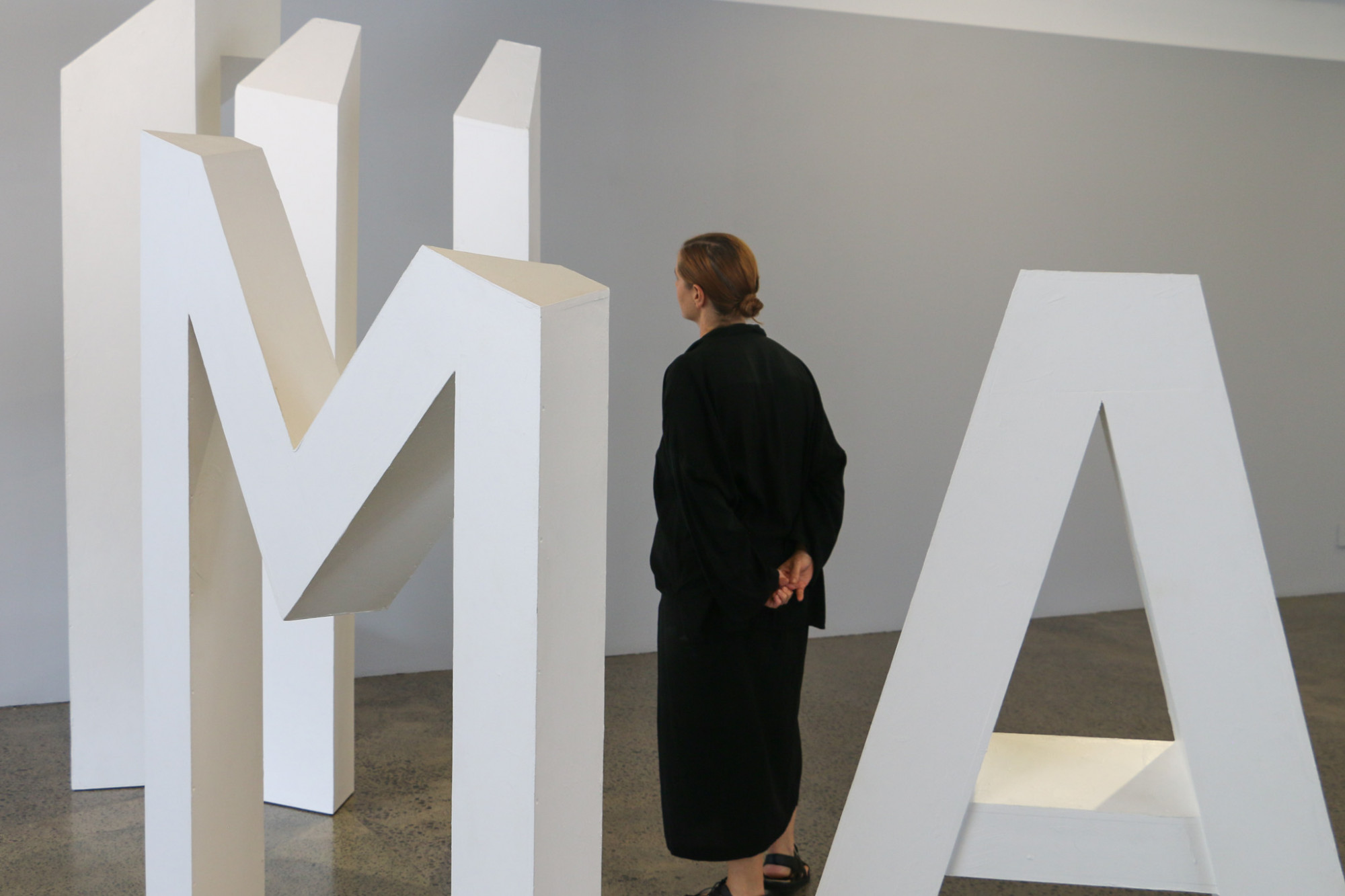 Image of part of a large white sculpture which spells out 'I AM HE', but only several of the letters are visible at this angle, a person is looking up at the top of the sculpture, pictured from behind.