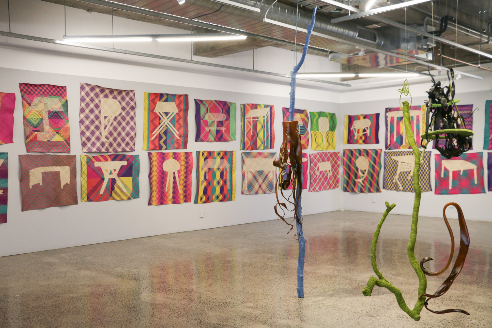 An installation of mats is hung on the wall and three sculptures made with seaweed are hung from the ceiling and one from a drift wood pole are in the foreground.