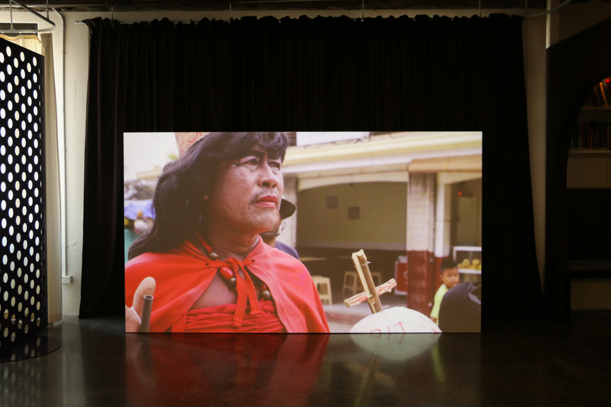 A projected film, capturing a film still of a man dressed in a costume including a red cape, lipstick and a wig, holding a religious cross.