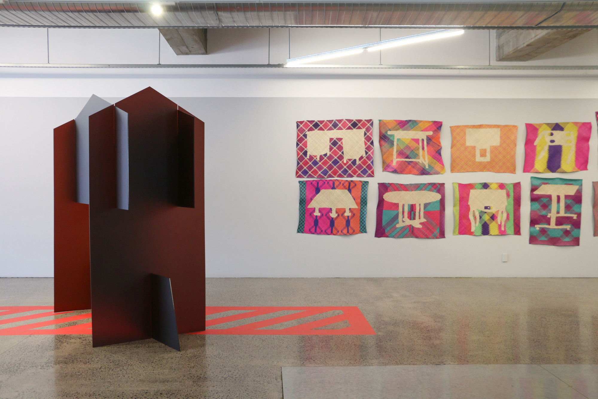 In the gallery to the left is a large perspex sculpture of flat pack design, resembling a Pā waharoa or gateway, but the top part of the structure obscures entry. Red floor vinyl maps out a path underneath the structure but has red horizontal lines running through it. Some colourful woven mats can bee seen on the wall behind.
