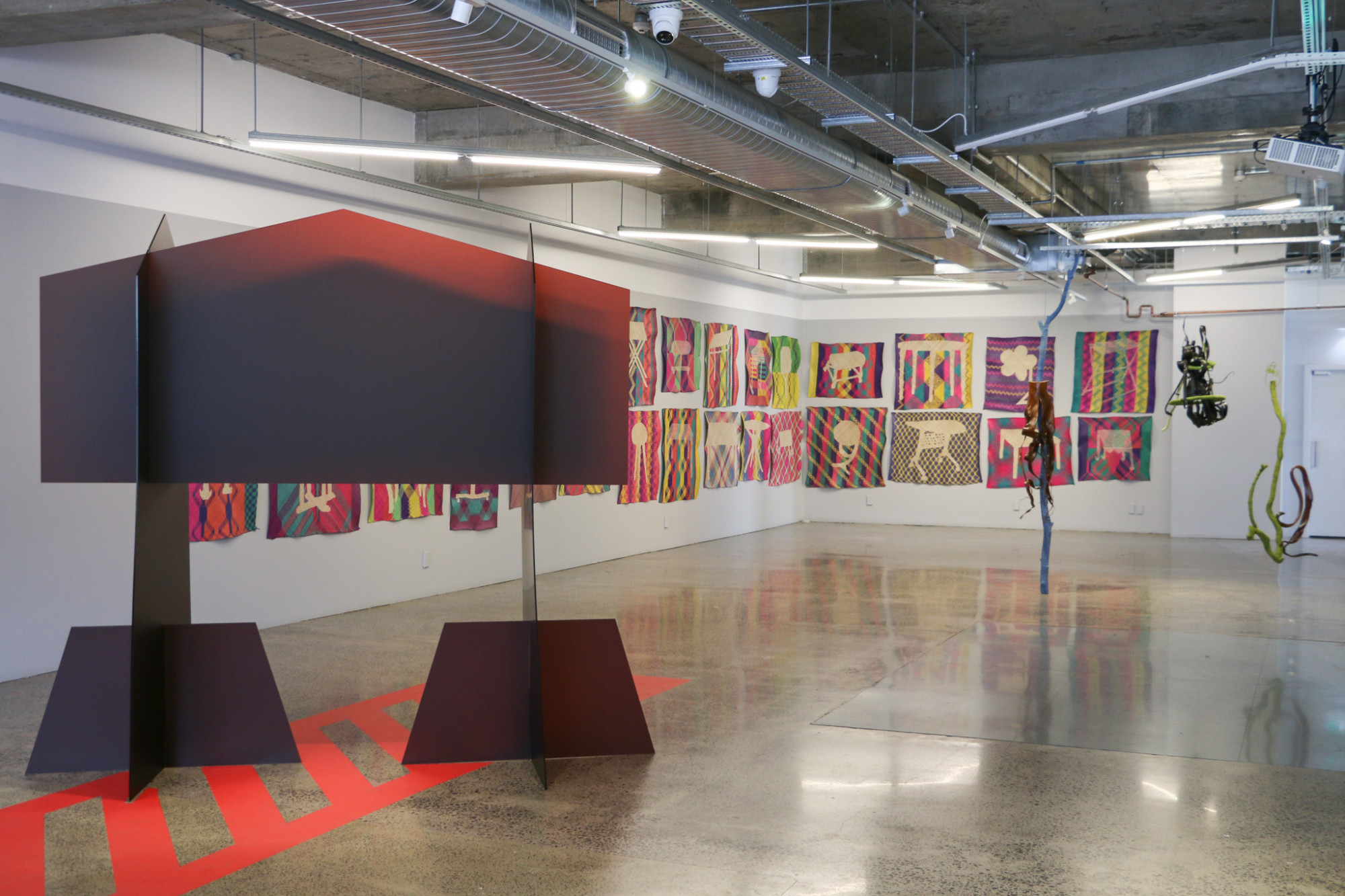 A gallery room with a large perspex structure with floor vinyl underneath, a series of mats hung on the walls and suspended sculptures at the back of the gallery.