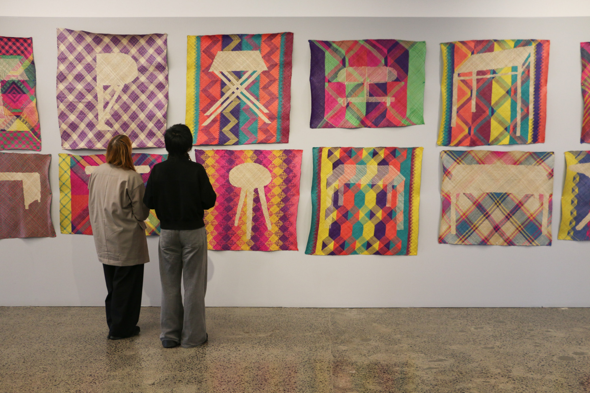Two people stand in front of a wall which has an installation of woven mats hung in two rows. Each mat is different with bright coloured patterned backgrounds and each has a silhouette of different style of table.