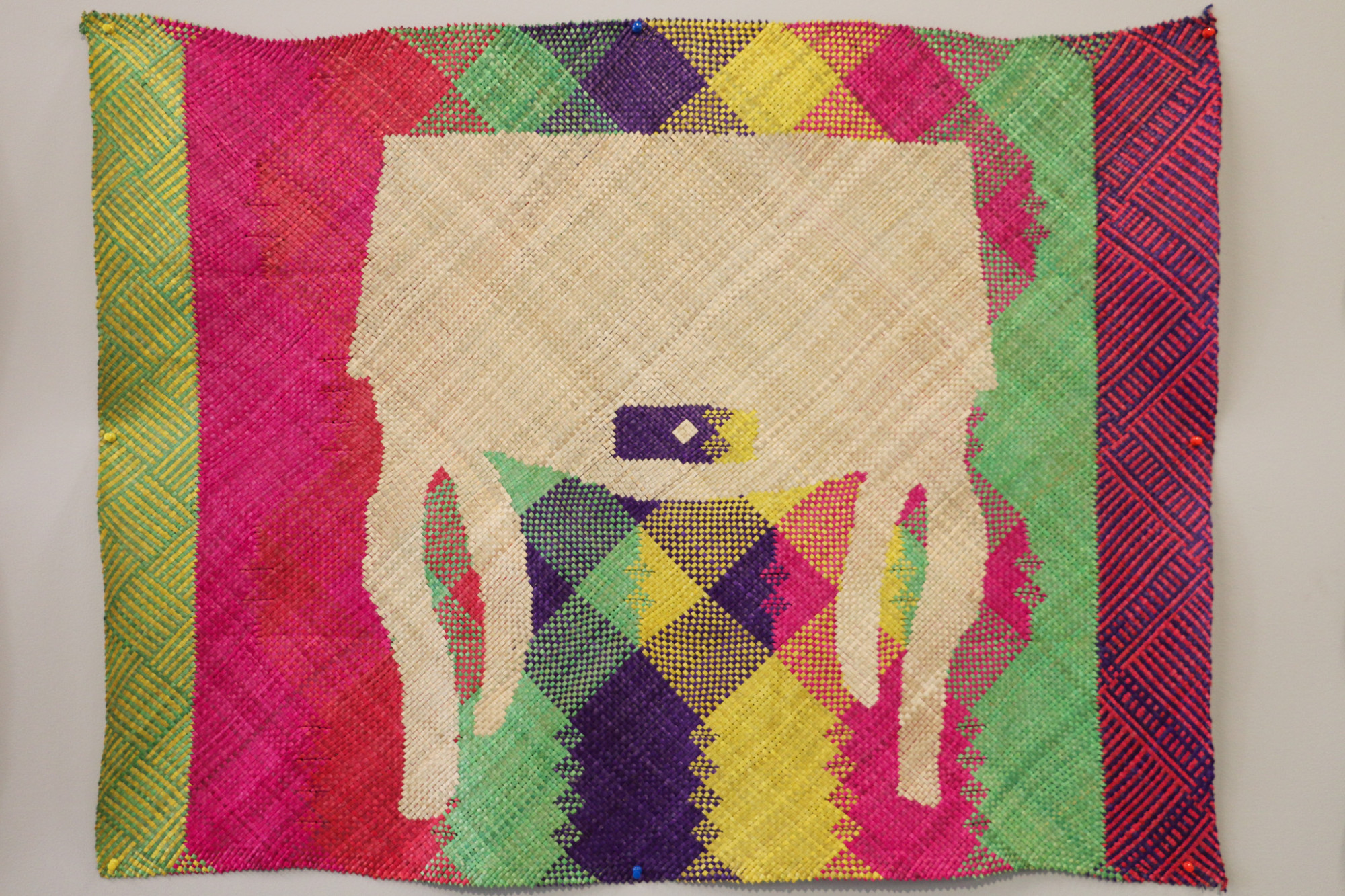 A installation detail of a single woven mat hung on the wall, which has silhouette of a desk in the middle with vertical stripes of bright colours which intersect as a grid pattern in the middle.