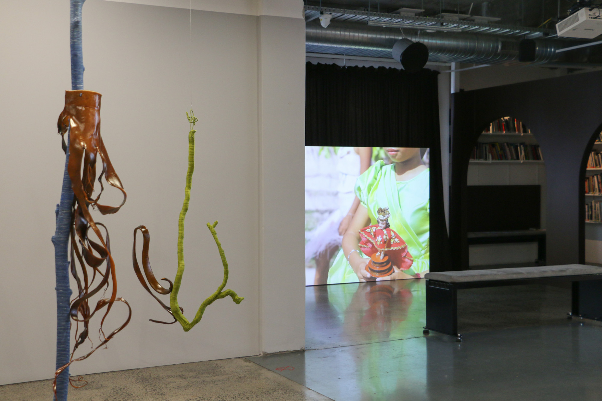 Two sculptures on the left are organic forms of seaweed and driftwood coated in coloured cord are suspended, the right there is a film projection featuring a child holding a religious statue obscured by a gallery wall.