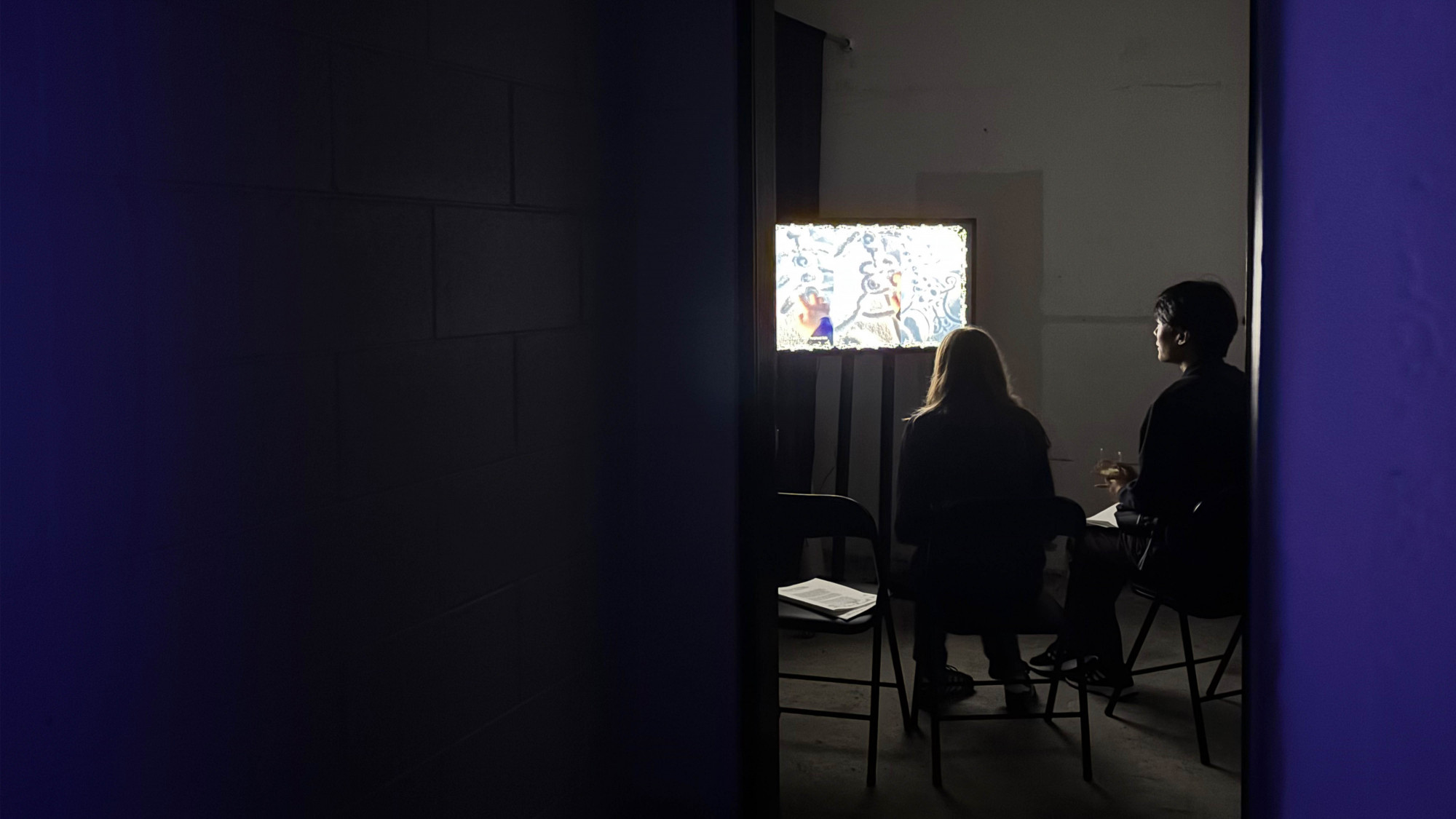 Two person sitting on black chairs in a small space watching a video on a tv screen