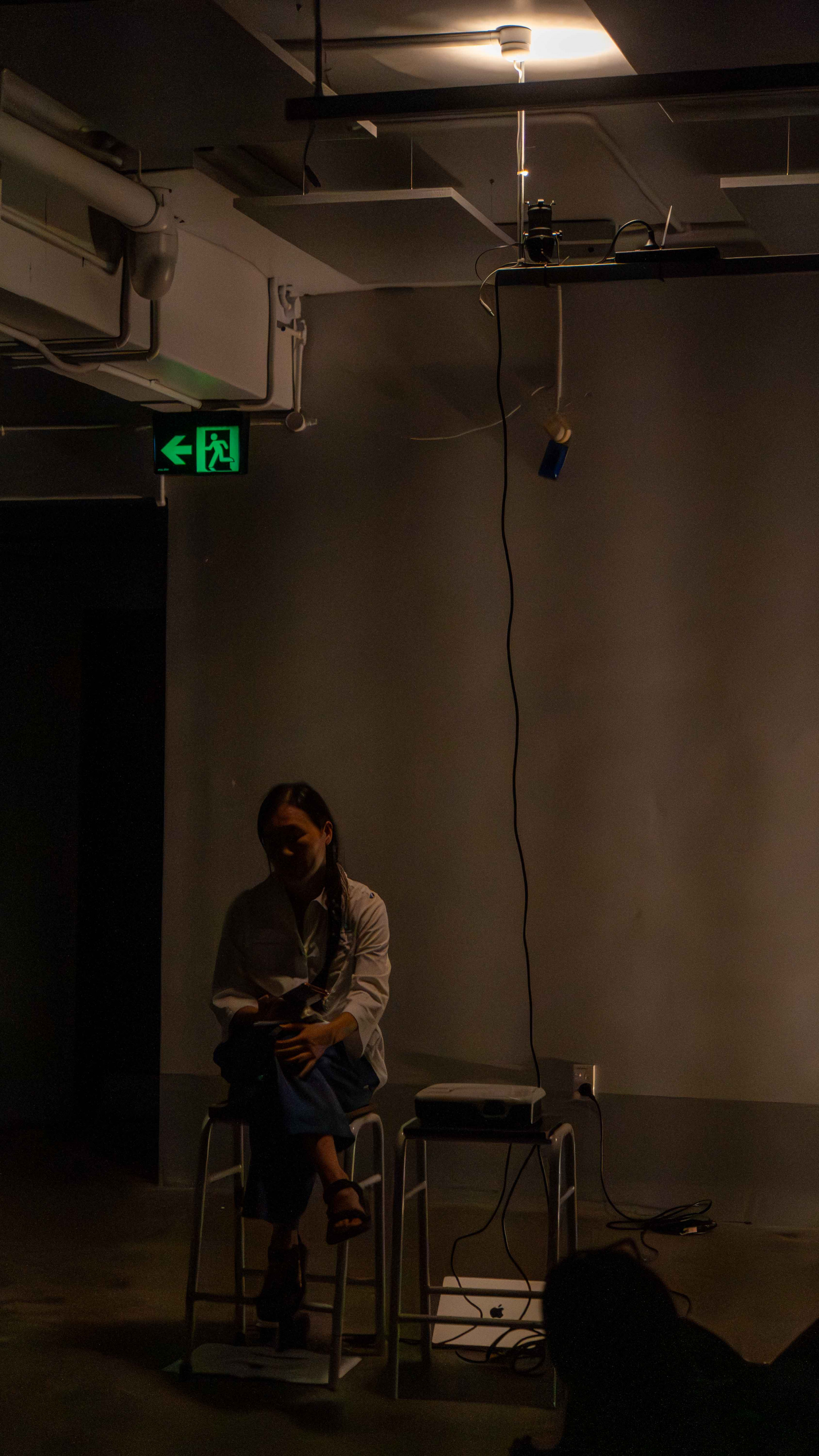 In a dark room with warm yellow light, a woman in white shirt sitting on a stool cross-legged