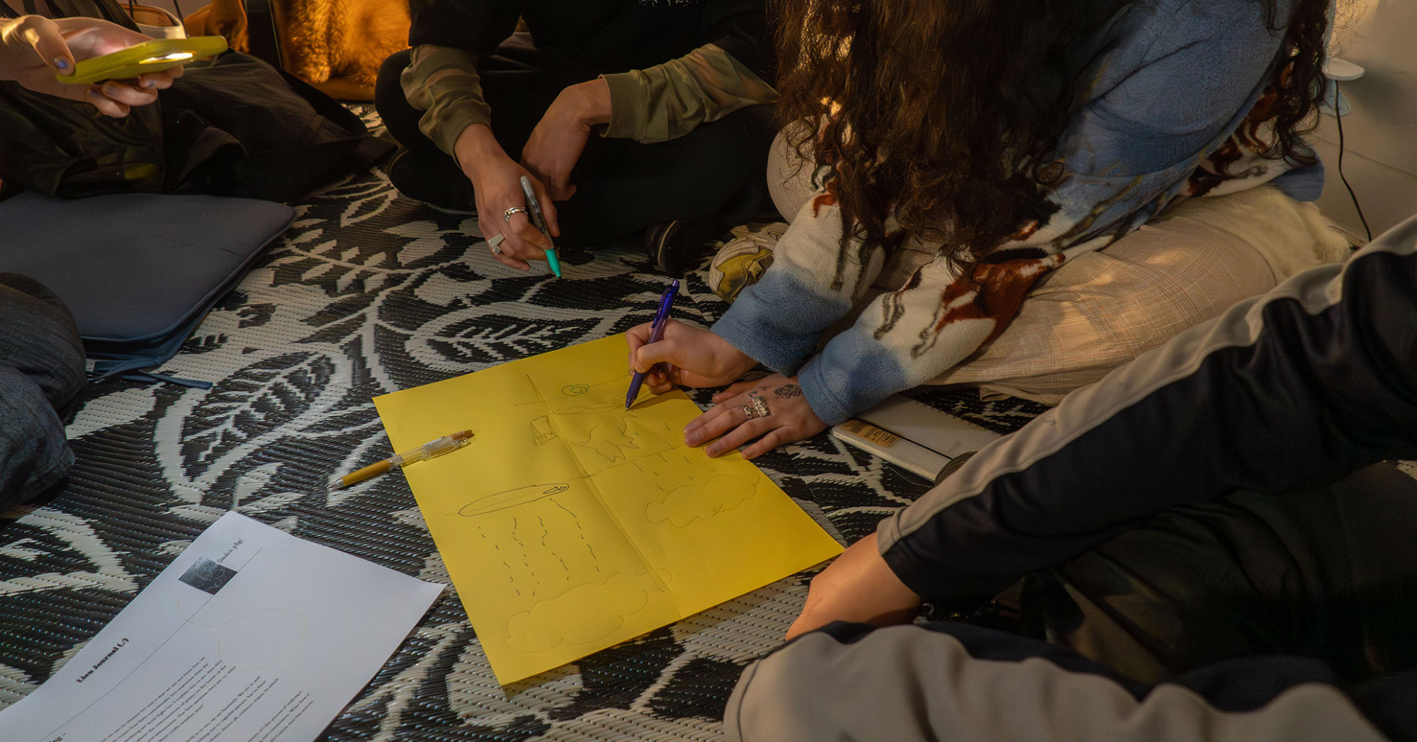 A few hands collectively drawing simple line raindrops and clouds on a piece of paper